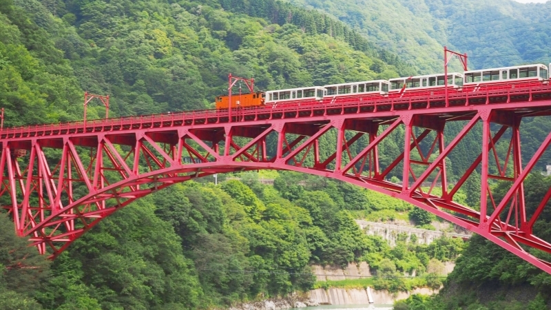 A scenic train crosses the red Kurobe Gorge Bridge in Japan’s lush mountains.