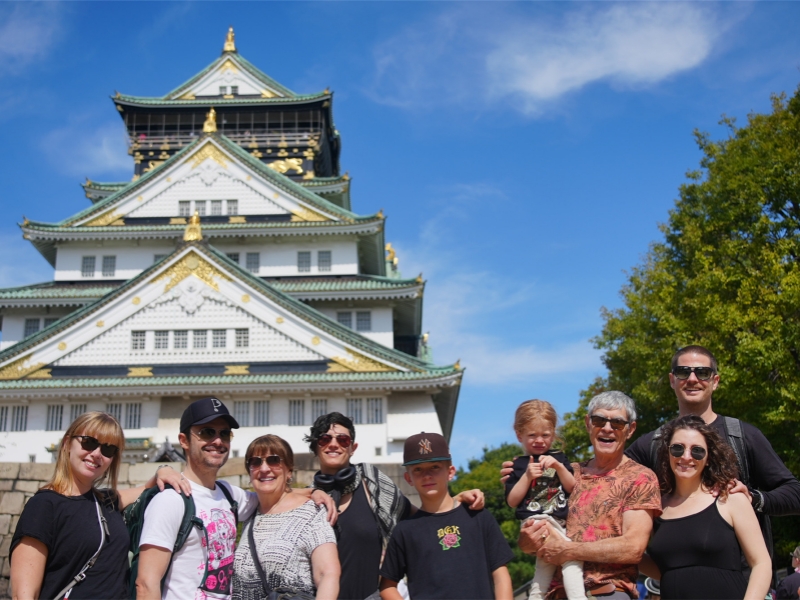 A happy multigenerational family pose for a photo in front of the iconic Osaka Castle on a sunny day.