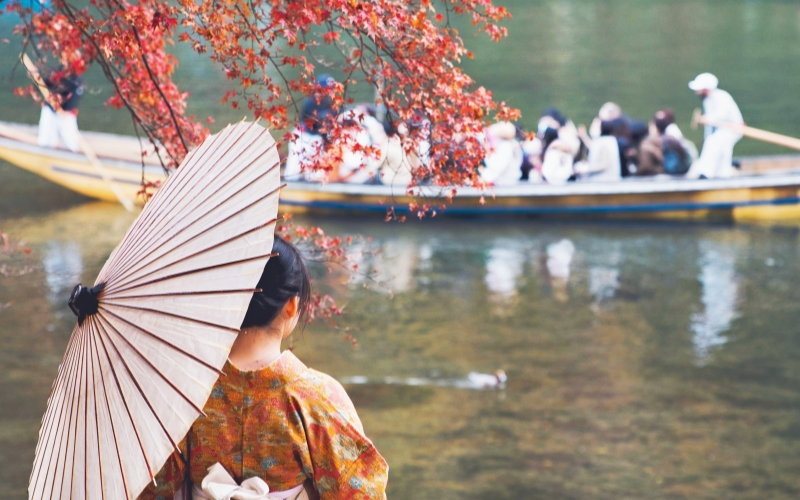 A woman in kimono enjoys the Arashiyama autumn view with red maple leaves and peaceful river boat rides in Kyoto.
