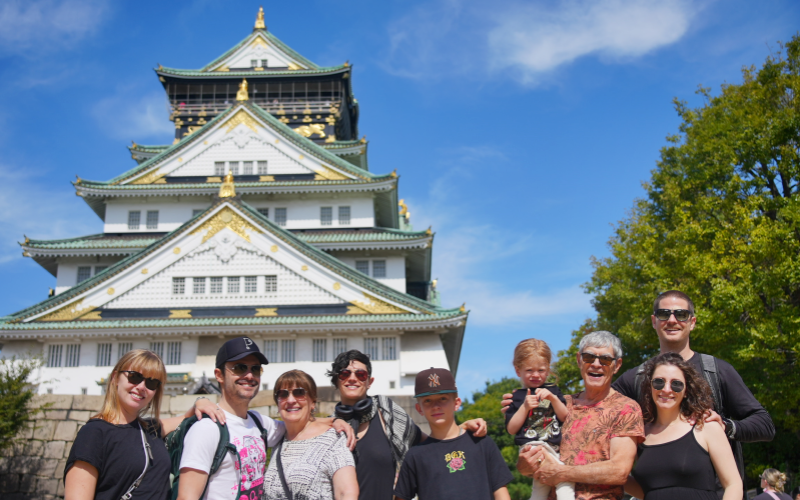 A cheerful family poses together in front of Osaka Castle on a bright, sunny day.
