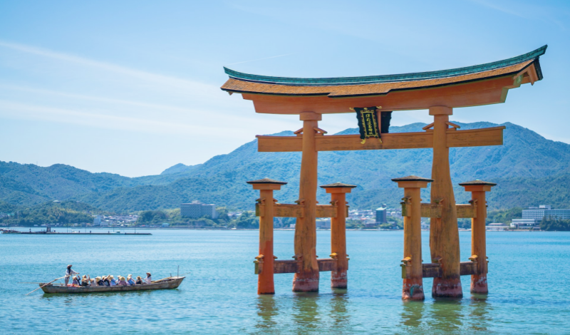 A traditional boat glides past the iconic floating torii of Miyajima on a clear, sunny day.