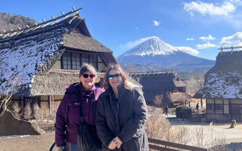 Clear blue skies and incredible visibility of Mount Fuji behind the snow-dusted traditional thatched-roof houses of Hakone Minshuku Village on a sunny day.