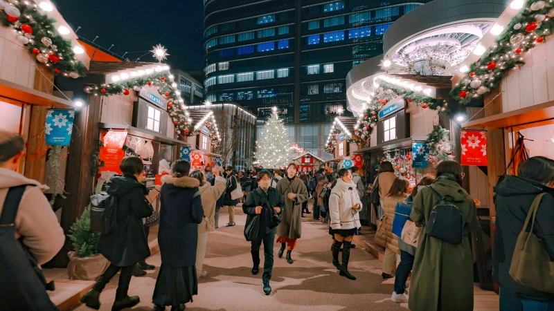 Festive crowds stroll past beautifully decorated, European-style wooden stalls toward a glowing holiday tree at the vibrant Azabudai Hills Christmas Market.