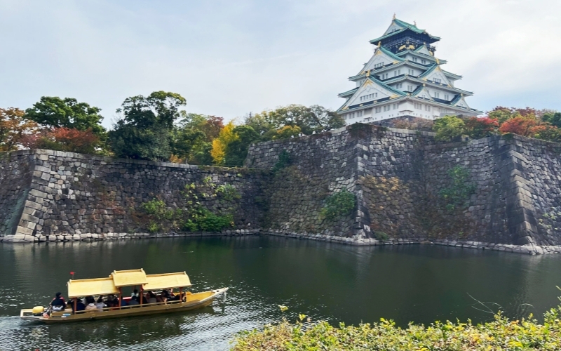 A traditional golden boat tours the moat of Osaka Castle during a mild and cloudy day typical of October weather in Osaka.
