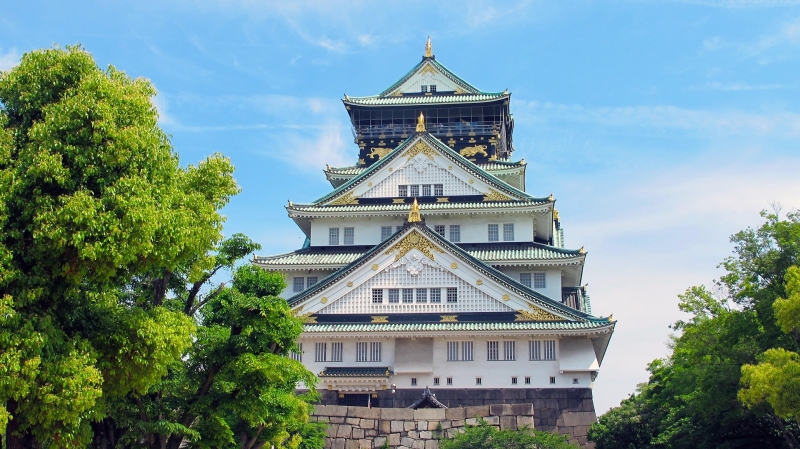Osaka Castle stands grandly under a clear blue sky, surrounded by lush June foliage during a sunny, warm early summer day.