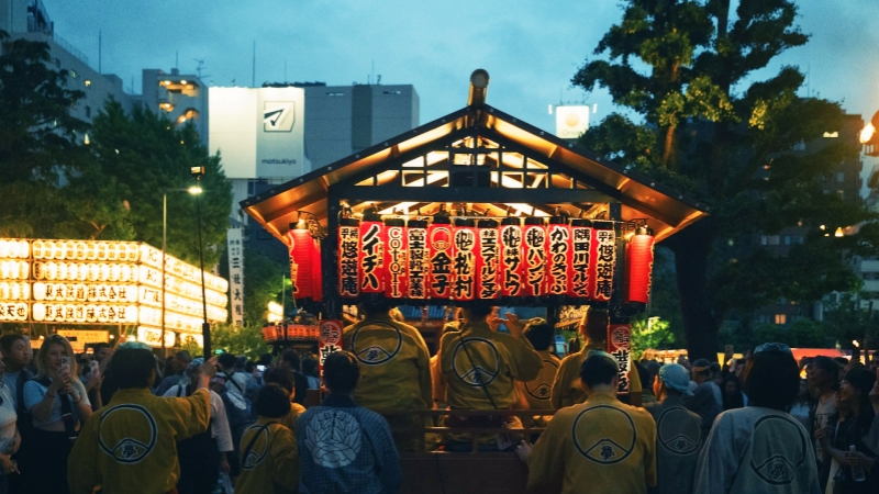 Under dusk skies, a crowd in traditional festival coats gathers around a brightly illuminated lantern stage in Asakusa, Tokyo.