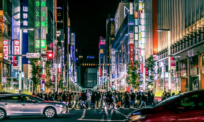 Bustling night in Tokyo's Ginza district, filled with people, cars, and illuminated neon signs.